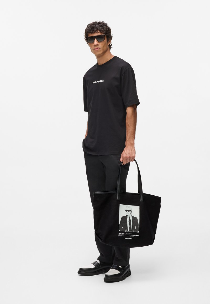 Crna torba s fotografijom figure koja nosi naočale, kožne remenčiće i mekanu teksturu. Model nosi crnu majicu i hlače.