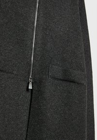 Dunkelgraue Strickjacke mit einem silbernen Reißverschluss, zwei aufgenähten Taschen und einem strukturierten Stoff für zusätzliche Tiefe.