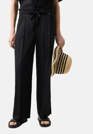 Persoon die losse zwarte broek met koord draagt, zwarte sandalen, en een beige strohoed met zwarte strepen vasthoudt in de rechterhand.