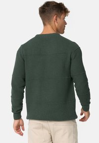 Pull en maille texturée verte avec un col rond, présentant des sections côtelées horizontales et des poignets ajustés, porté sur un pantalon de couleur claire.