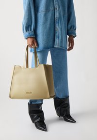 Sac fourre-tout beige au design structuré et aux poign�ées doubles, arborant un logo "REPLAY" bien visible à l'avant. Présenté avec une tenue en denim bleu.
