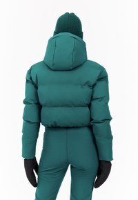 Groene pufferjack met een hoge kraag en capuchon, met een gequilt ontwerp. Gecombineerd met zwarte handschoenen en bijpassende groene broek.