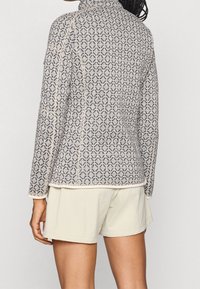 Osoba v béžovém a tmavě šedém vzorovaném svetru s dlouhým rukávem a vysokým výstřihem, v kombinaci se světle béžovými šortkami, zobrazená ze zad.