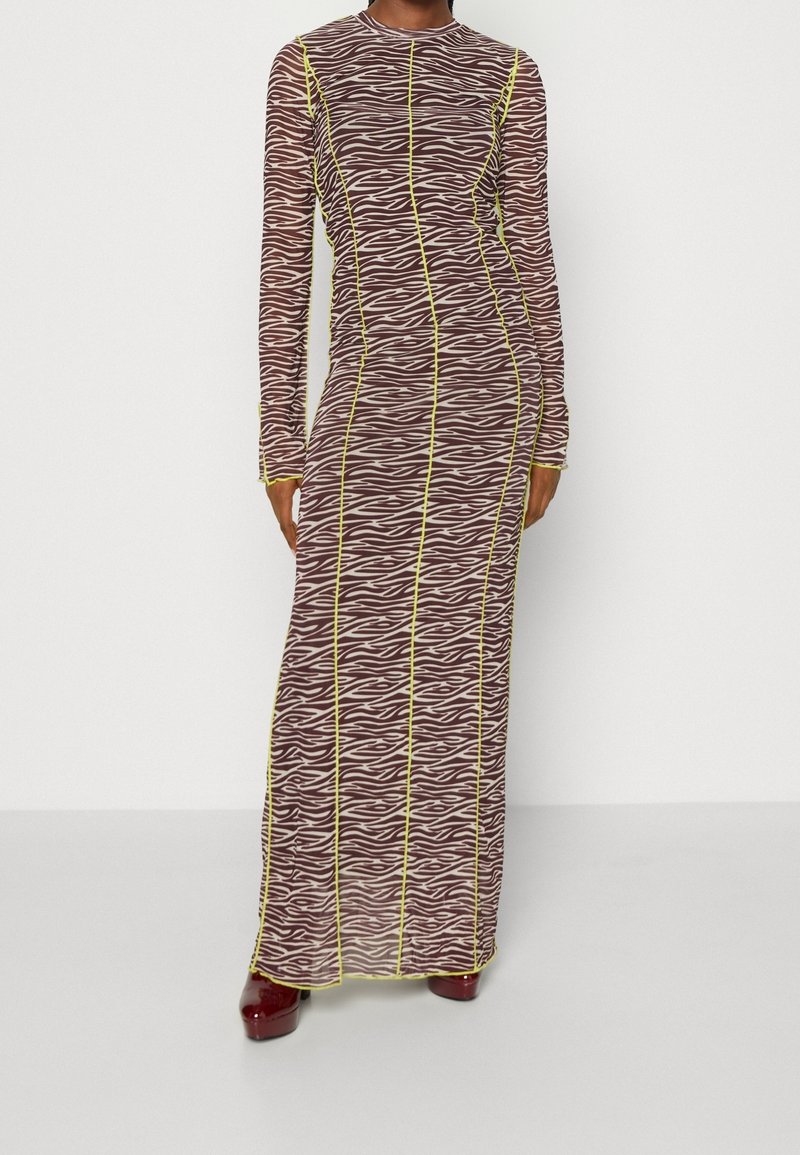 Femme portant une robe longue à manches longues avec des rayures zébrées marron et crème et des coutures verticales jaunes, associée à des chaussures bordeaux.