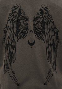 Μαύρα λεπτομερή φτερά αγγέλου με ημισεληνοειδείς και αστρικά σύμβολα τοποθετημένα κάθετα μεταξύ τους πάνω σε σκούρο ύφασμα.