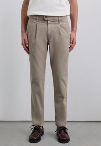 Pantalones de algodón beige con pliegues frontales, cierre de botón y piernas ajustadas. Llevados con zapatos de cordones marrón oscuro.
