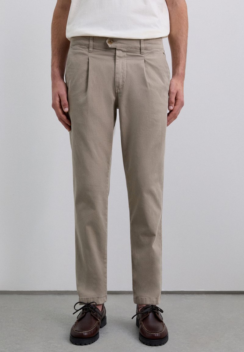 Pantalones de algodón beige con pliegues frontales, cierre de botón y piernas ajustadas. Llevados con zapatos de cordones marrón oscuro.