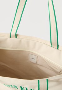 Μπεζ τσάντα tote Calvin Klein με λαβές με πράσινες ρίγες και εσωτερική τσέπη με φερμουάρ, εμφανίζει ετικέτα υφάσματος και εξωτερικό κείμενο με την επωνυμία.