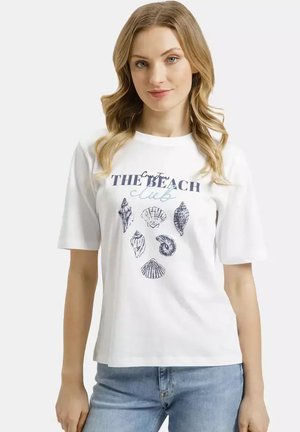 Młoda kobieta z falującymi blond włosami, ubrana w białą koszulkę z ilustracjami muszelek i napisem "The Beach Club", oraz niebieskie dżinsy.