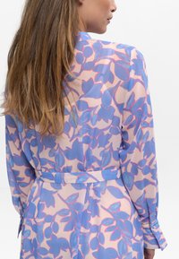 Blouse en tissu léger et translucide avec un motif floral bleu et rose. Manches longues avec poignets boutonnés et taille cintrée.