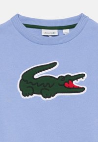 Felpa azzurra chiara con logo di coccodrillo verde con accenti rossi, contornato di bianco, e colletto a girocollo a coste.