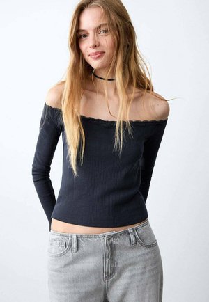 Bluză neagră cu dungi, decoltată pe umeri cu guler ondulat, mâneci lungi și croială mulată, asortată cu blugi gri deschis, talie înaltă.