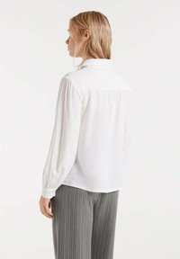 Mujer rubia de perfil izquierdo, vistiendo una blusa blanca de manga larga con mangas ajustadas y pantalones grises a rayas contra un fondo liso.