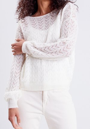 Femme portant un pull en maille blanc avec un motif chevron et un pantalon blanc, debout devant un fond clair uni.