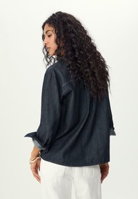 Camicia in denim scuro con vestibilità rilassata, polsini con bottoni e maniche arrotolate. Presenta un colletto classico e un sottile dettaglio di cucitura sul retro.