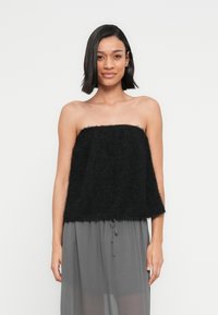 Νέα γυναίκα με σκούρα μαλλιά μήκους ώμων φοράει ένα μαύρο τοπ χωρίς ράντες από μαλακό ύφασμα και φαρδιά γκρι παντελόνια μπροστά σε απλό φόντο.