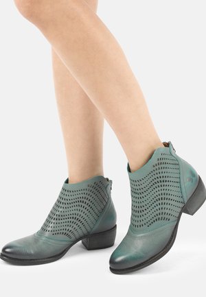 Ankelhøje støvler i blågrønt læder med perforerede bølgemønstre, båret på krydsede bare ben.