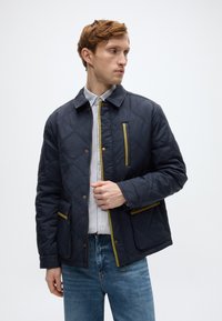 Ναυτικό μπουφάν με ραφές σε σχέδιο διαμαντιού, πράσινες λεπτομέρειες, κουμπιά με πάτημα και πλαϊνές τσέπες. Φορεμένο πάνω από ριγέ πουκάμισο με μπλε τζιν.