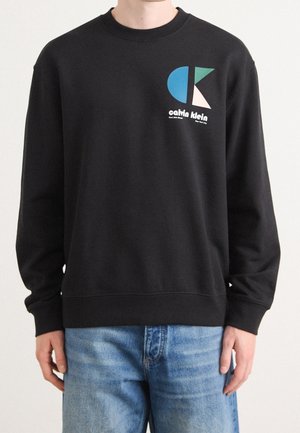 Czarna bluza z kolorowym geometrycznym logo Calvin Klein na lewej piersi, noszona z jasnoniebieskimi dżinsami.