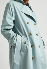 Jasnoniebieski dwurzędowy trencz z dużymi brązowymi guzikami, bocznymi kieszeniami i paskiem. Wykonany z gładkiej tkaniny o dopasowanym kroju.