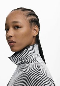 Pull en maille noir et blanc à col montant. Modèle avec des cheveux tressés, un teint neutre et des traits du visage marqués. Fond blanc.