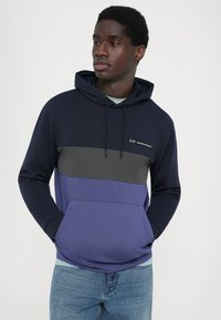 Sweatshirt com capuz azul marinho e roxo, com uma faixa horizontal cinza, bolsa frontal e capuz com cordão. O tecido parece suave e elástico.