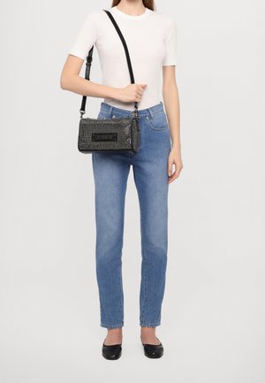 Bolso negro adornado con un acabado texturizado y correa ajustable, que presenta una placa con logotipo y pequeños studs redondos. Lleva puesto con jeans azules.