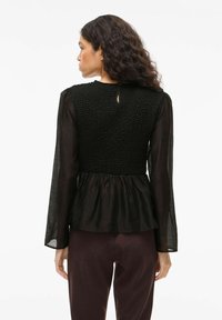 Top nero a peplo con corpetto lavorato a maglia testurizzato, maniche lunghe trasparenti e dettaglio a occhiello sul retro. Progettato con una vestibilità ampia e rilassata.