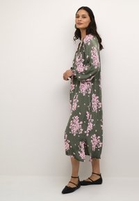 Rochie fluidă de măsline cu un model floral roz, mâneci lungi și o lungime medie. Asortată cu pantofi negri fără toc, împodobiți cu bretele.