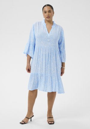 Kobieta stoi ubrana w jasno niebieską sukienkę do kolan z subtelnym kwiatowym wzorem, rękawami 3/4 typu dzwon, z marszczonym dekoltem i czarnymi sandałami na obcasie.