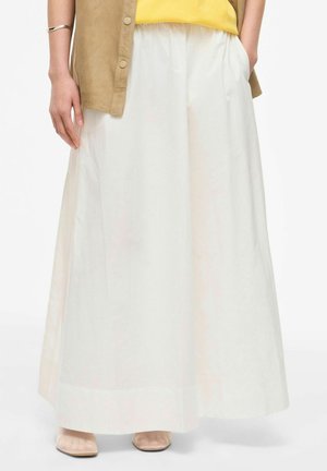 Nainen, jolla on pitkä valkoinen hame, keltainen toppi, beige avoin paita, beiget kengät ja hopeinen rannekoru, seisomassa käsi taskussa.