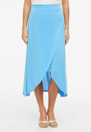 Femme portant une jupe mi-longue style portefeuille bleu clair et des sandales plates beiges à bout ouvert, debout devant un fond uni.