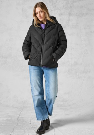 Doudoune noire avec capuche, arborant un motif en chevrons. Associée à un jean bleu clair et des bottes noires imposantes. Le modèle se tient dans un cadre neutre.
