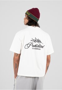 Hvit T-skjorte med kort erm som har svart tekst og et bladgrafikk på baksiden. Kombinert med en maroon lue med grønne og gule detaljer.