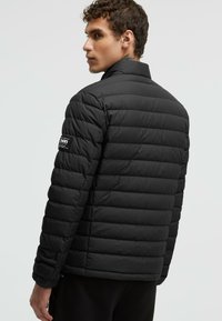 Black puffer jacket with a quilted design, high collar, and a logo patch on the left shoulder. Soft, textured fabric with a slim fit.