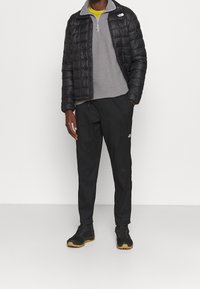 Svart puffjacka över en grå fleecepullover, tillsammans med svarta byxor och svarta sportskor med gummisula, med varumärkeslogotyper.