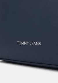 Marineblå syntetisk läderväska med präglad silver "TOMMY JEANS"-logotyp. Slät textur, elegant rektangulär form, minimalistisk design.