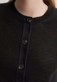Schwarzer Strickcardigan mit rundem Ausschnitt, geripptem Kragen und drei schwarzen Knöpfen an der Vorderseite, der eine strukturierte Oberfläche zeigt.