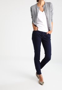 Šedý cardigan s knoflíky přes bílou košili s výstřihem do V, doplněný tmavě modrými džíny a stříbrnými balerínami. Hladké textury a přiléhavý design.