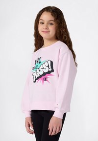 Chica con el cabello largo y rizado, vistiendo un sudadera rosa claro con el colorido texto "NYC Vibes? Yasss!" y pantalones negros, de pie contra un fondo blanco.