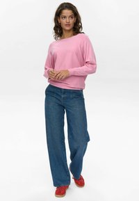 Camisa rosa de manga larga con corte relajado, combinada con unos vaqueros de mezclilla amplios en azul. El modelo lleva zapatillas rojas, mostrando un conjunto casual.