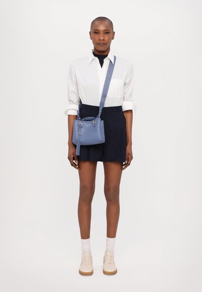 Borsa a tracolla in pelle blu con hardware argentato, design strutturato, tracolla regolabile e etichetta con logo, abbinata a una camicia bianca e pantaloni corti blu navy.