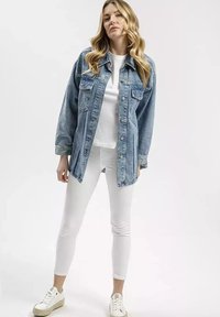 Jasnoniebieska dżinsowa kurtka o luźnym kroju, z dużymi kieszeniami z przodu, zapinana na guziki, noszona na białą koszulkę i białe dopasowane spodnie.