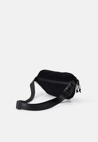 Svart corduroy midjeveske med glidelås, justerbar stropp og teksturert overflate. Har en kompakt, avrundet form.