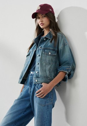 Junge Frau, die eine burgunderrote Kappe, eine Jeansjacke und hoch taillierte Jeans trägt, posiert mit einer Hand in der Tasche vor einem einfarbigen Hintergrund.