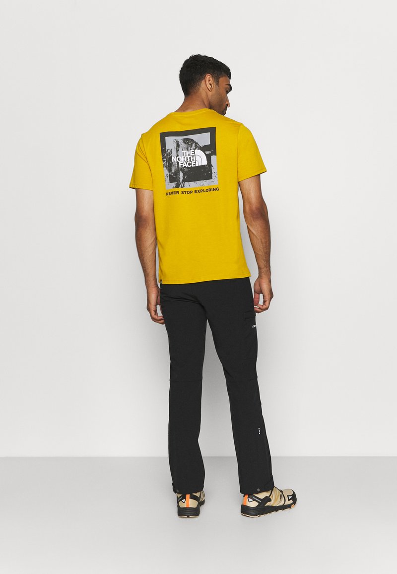 Gele katoenen T-shirt met een zwart grafisch ontwerp op de achterkant. Gecombineerd met zwarte broek en beige schoenen met oranje accenten.