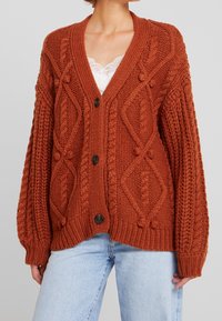 Rdzawoczerwony sweter z dzianiny z teksturowanym wzorem, dekoltem w kształcie litery V i brązowymi guzikami. Rękawy są szerokie z ściągaczami.