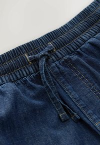 Cintura de pantalones vaqueros con banda elástica y cordón ajustable, con costuras visibles y detalle de bolsillo frontal.