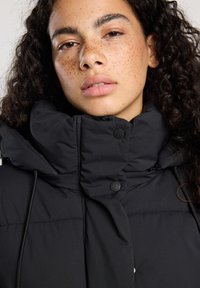 Doudoune noire avec un col haut, des boutons et une finition texturée. Le design comprend un cordon de serrage et une fermeture éclair.
