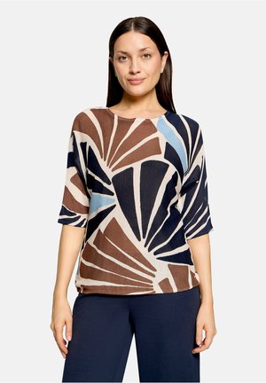 Vrouw met stijl donker haar die een top met korte mouwen draagt met bruine, marineblauwe en lichtblauwe abstracte waaierpatronen en marineblauwe broek.